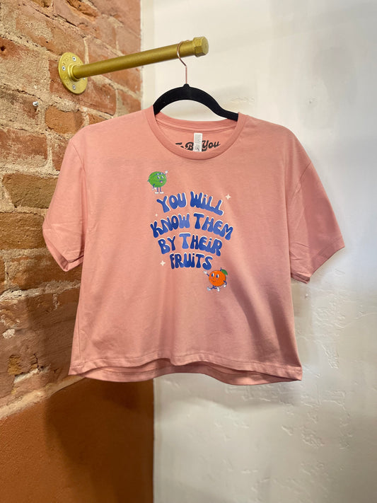 This cropped t-shirt features a front graphic depicting cartoon fruit and the text “You will know them by their Fruits”. Colorful graphic on a cropped pink tee. 